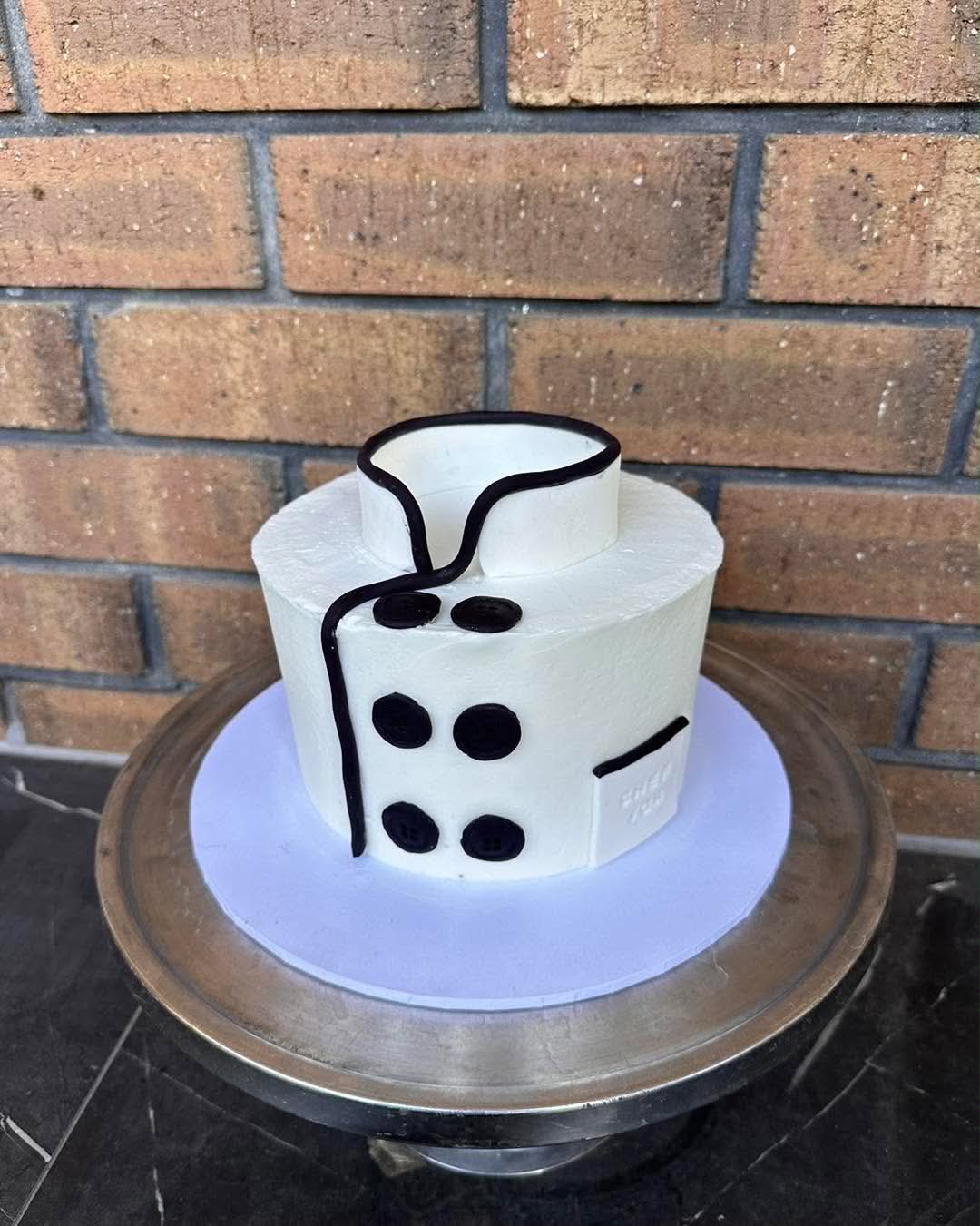 White pearl dot Baat Pakki engagement cake with gold acrylic ring topper on silver cake stand surrounded by pebbles and greenery